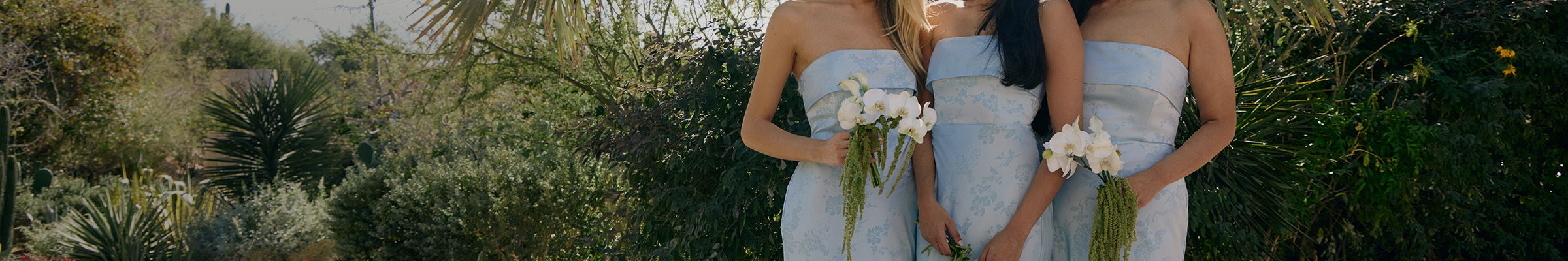 Three women in light blue dresses holding flowers outdoors.