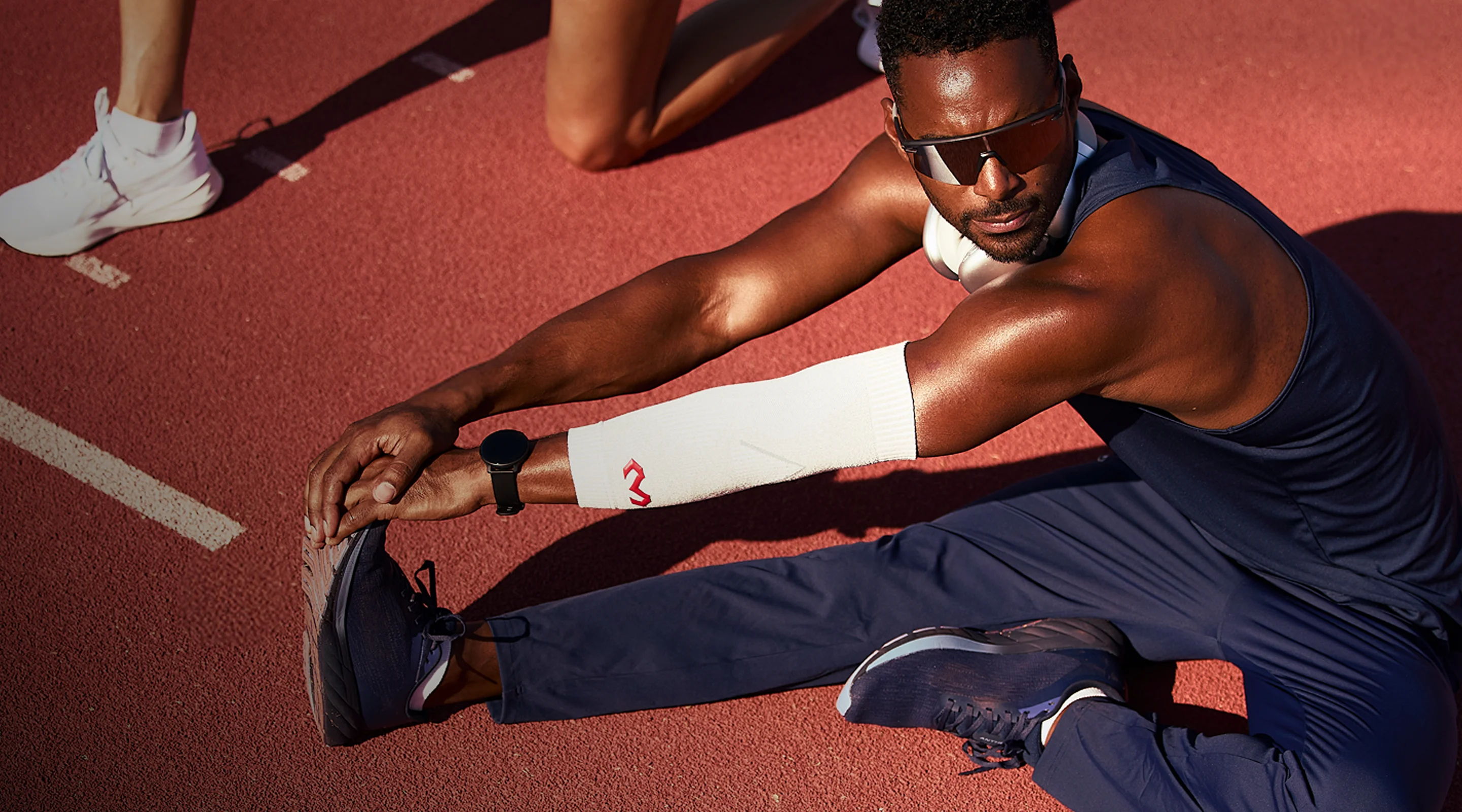 Athlete stretching on a track, wearing sports attire and sunglasses.