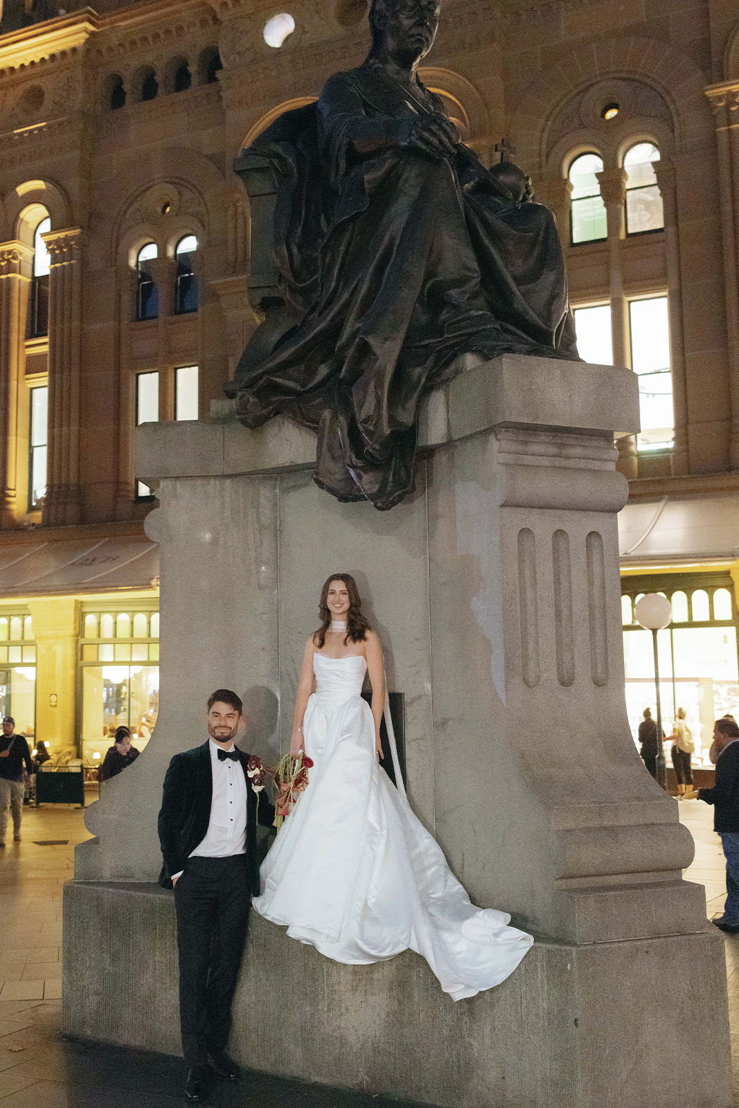 Bride and groom posing by a statue in a public space.