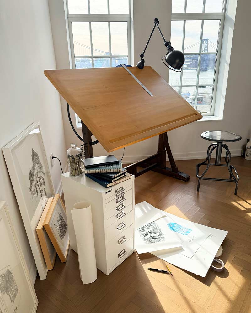 An artist's workspace with a drafting table, lamp, and scattered materials.