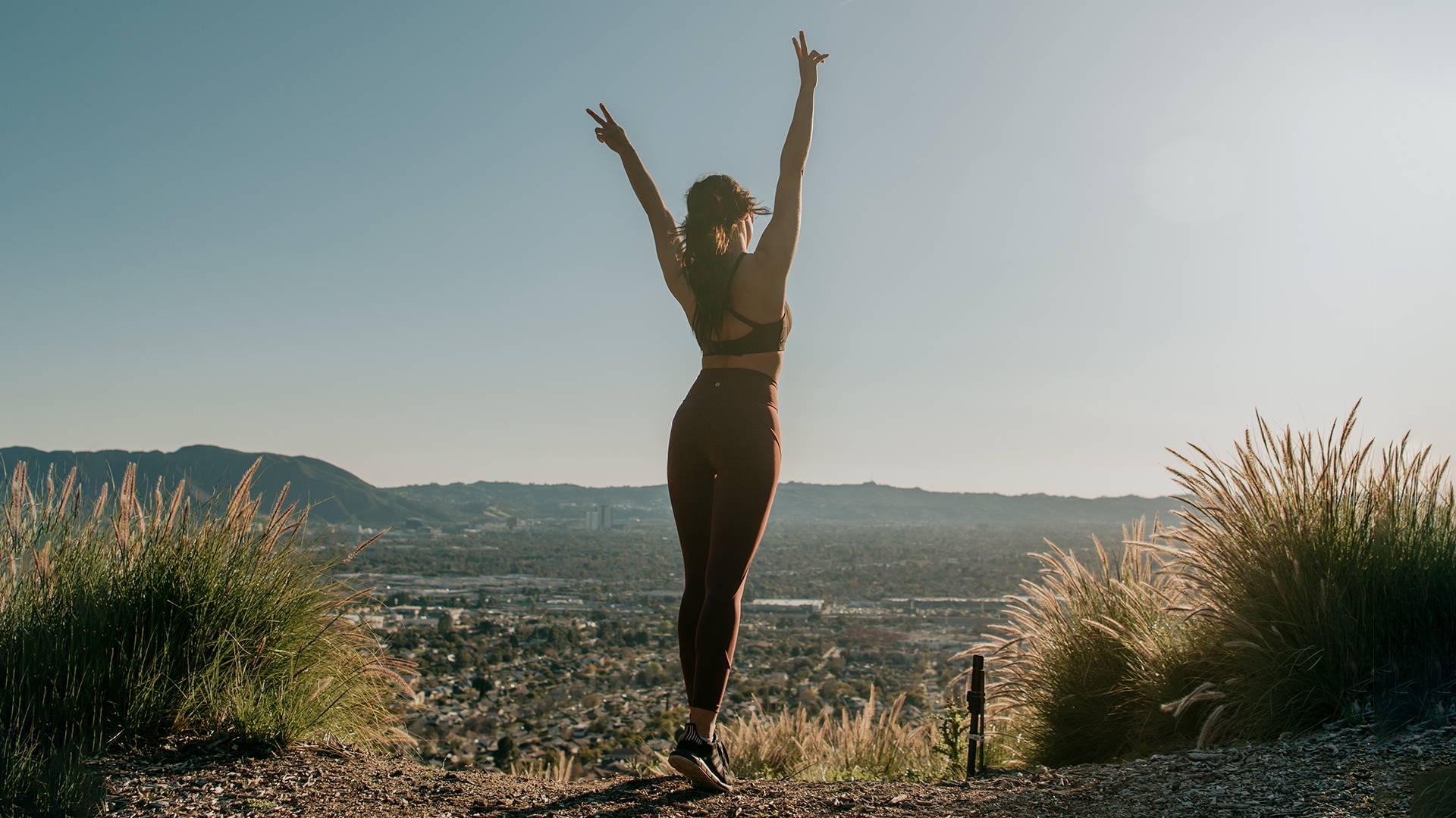 Woman in yoga attire doing two peace signs in the air over looking a large landscape.