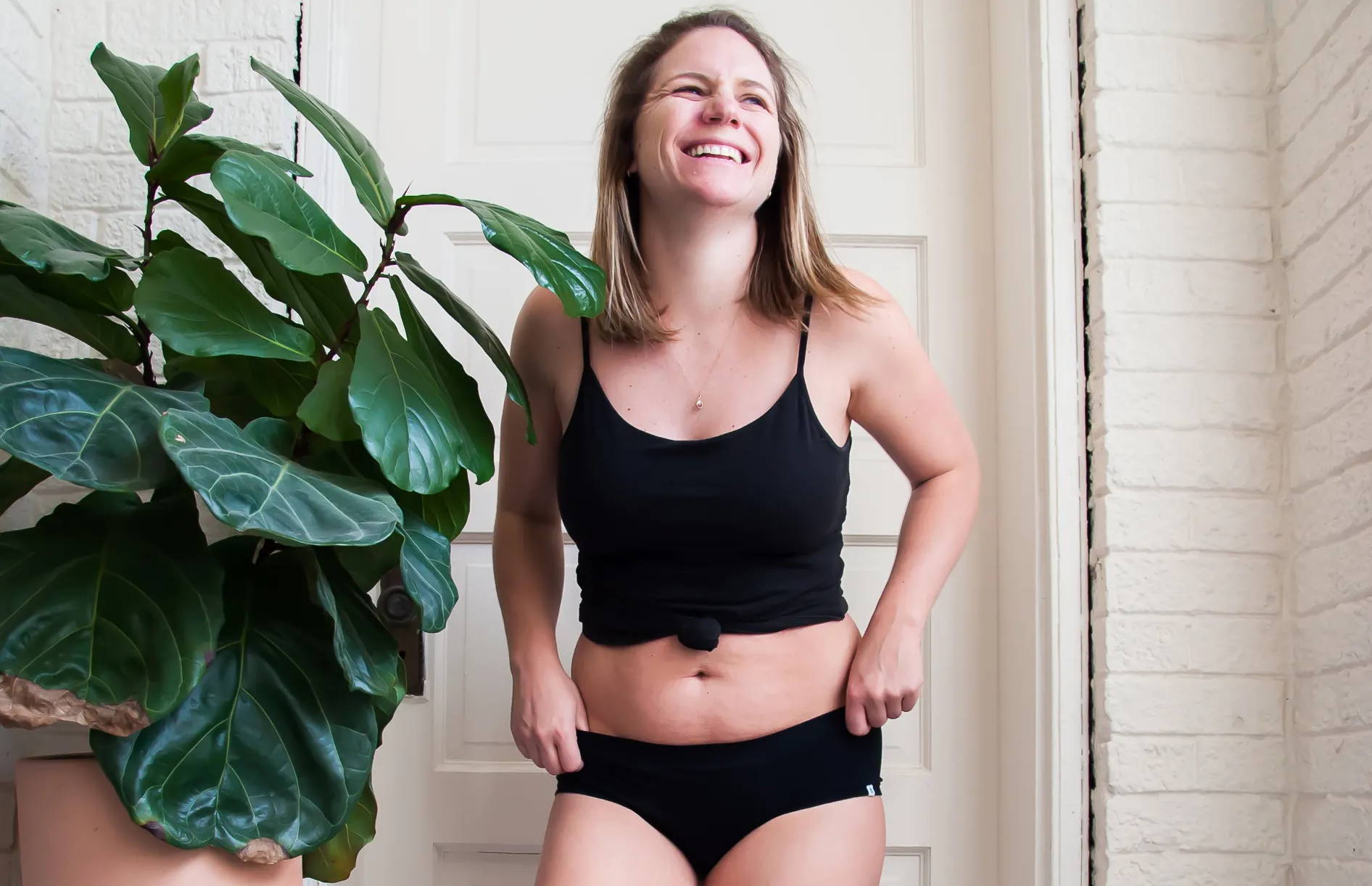 A woman stands next to a plant in WAMA hemp underwear and a tank top, smiling.