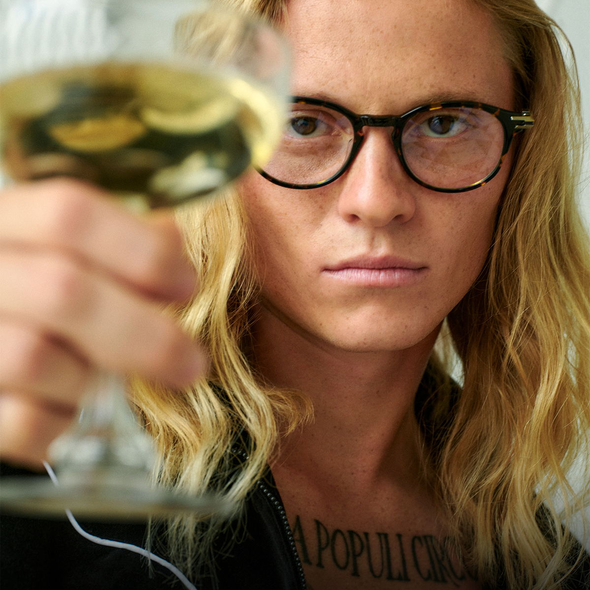 Person with long hair and glasses holding a glass of wine.