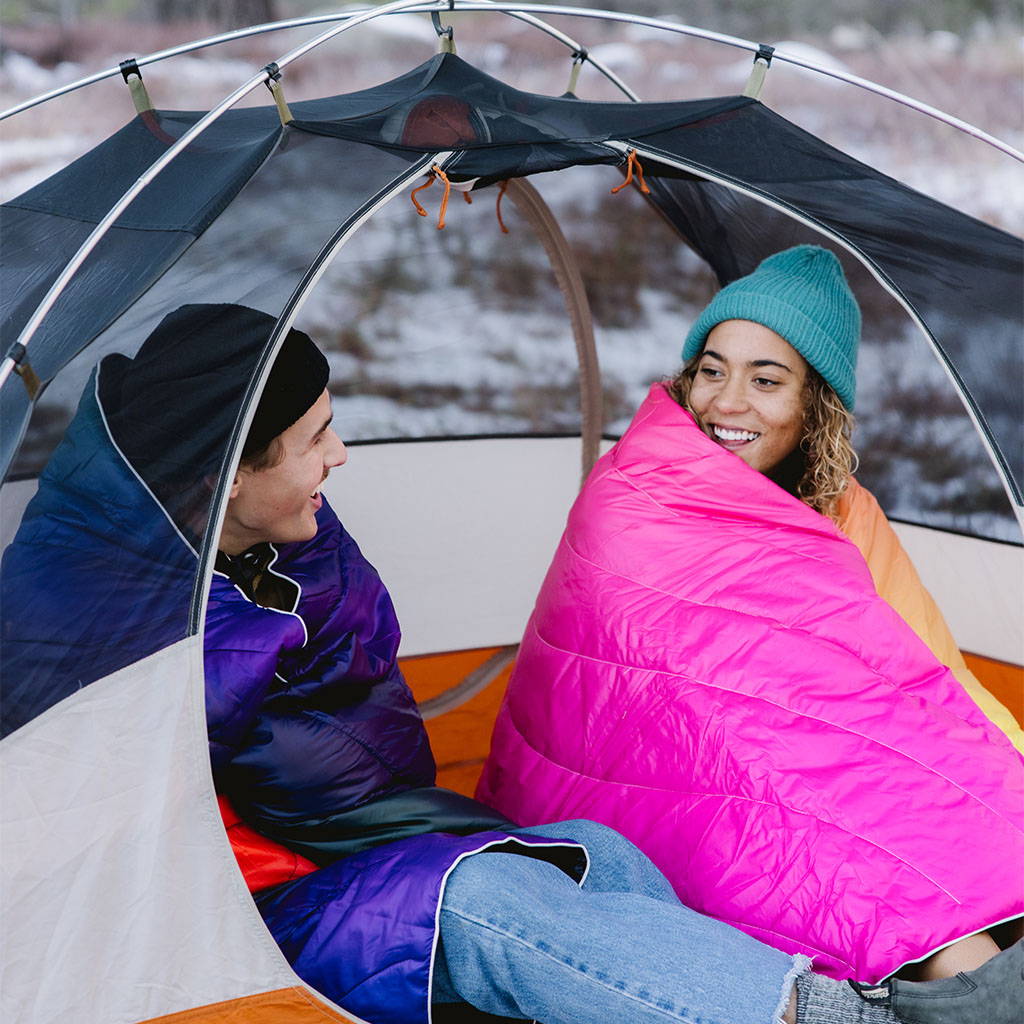 Two friends sitting and talking in a tent while wrapped in warm Rumpl blankets.