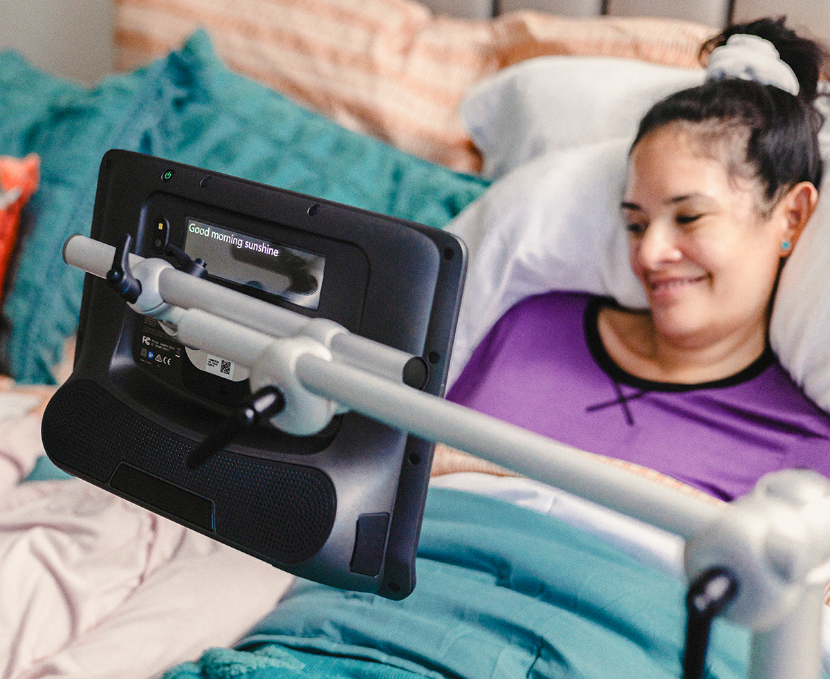 Floorstand mount for a Tobii Dynavox assistive communication device used by a woman in her bed