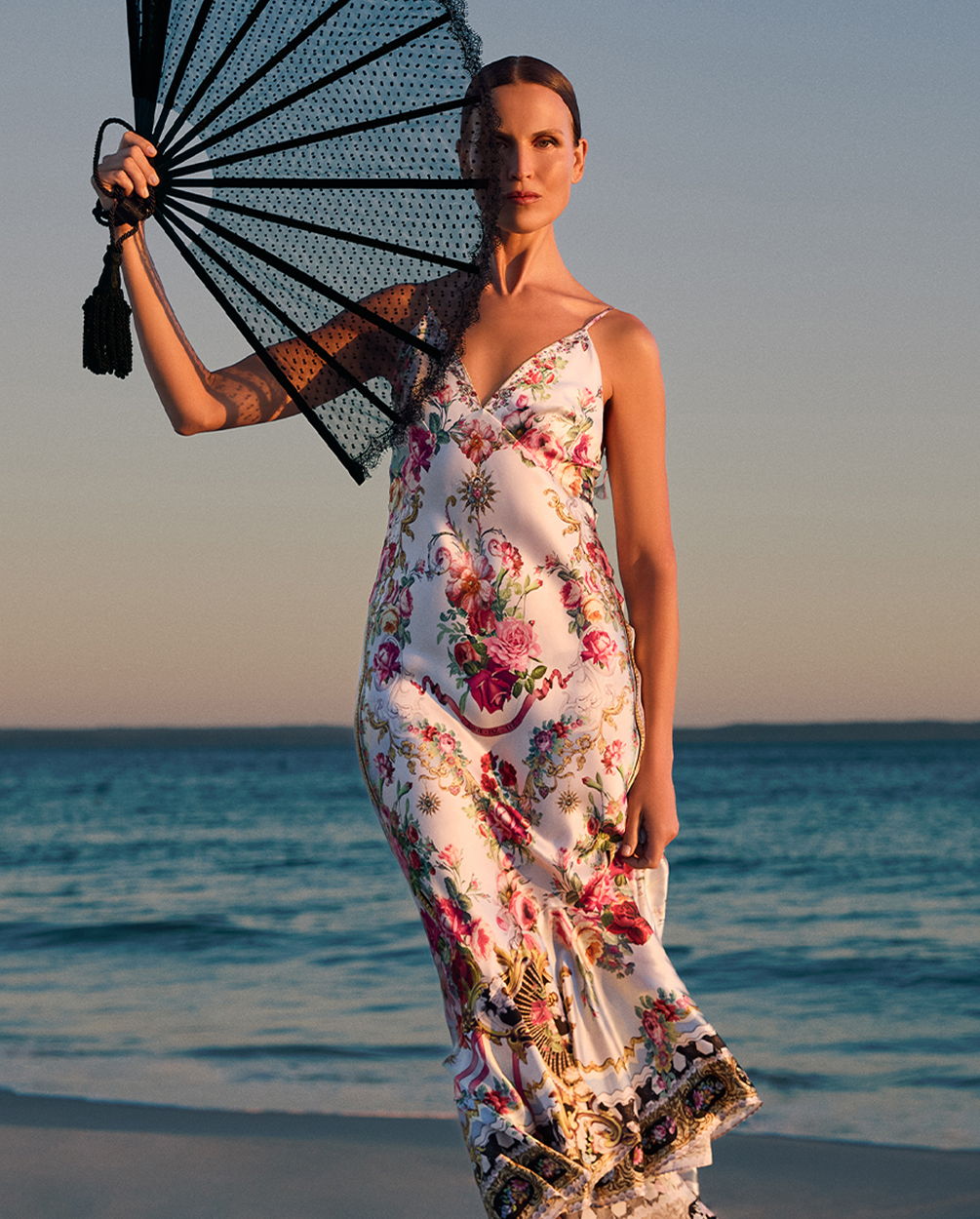 Woman in a floral dress holding a fan by the beach at sunset.