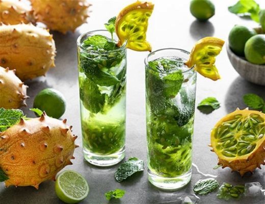 Two tall glasses of green mint drinks with fruit and limes around them.