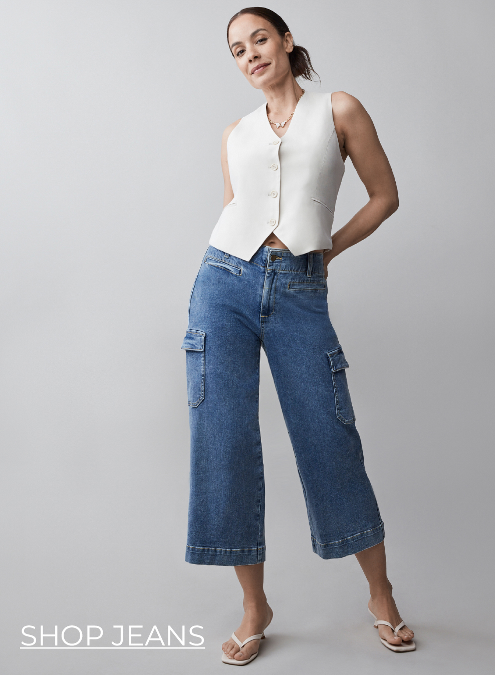 Woman wearing a white vest and blue cargo jeans, smiling against a gray background.