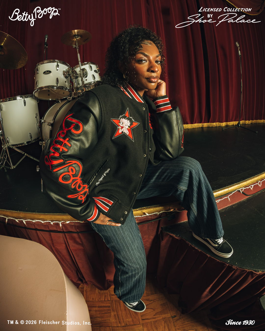 Person in a varsity jacket posing near a drum set on stage.
