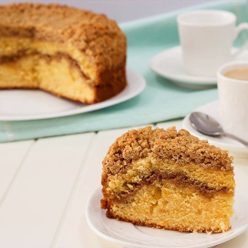 slice of coffee cake in the foreground with larger cake in background with mugs