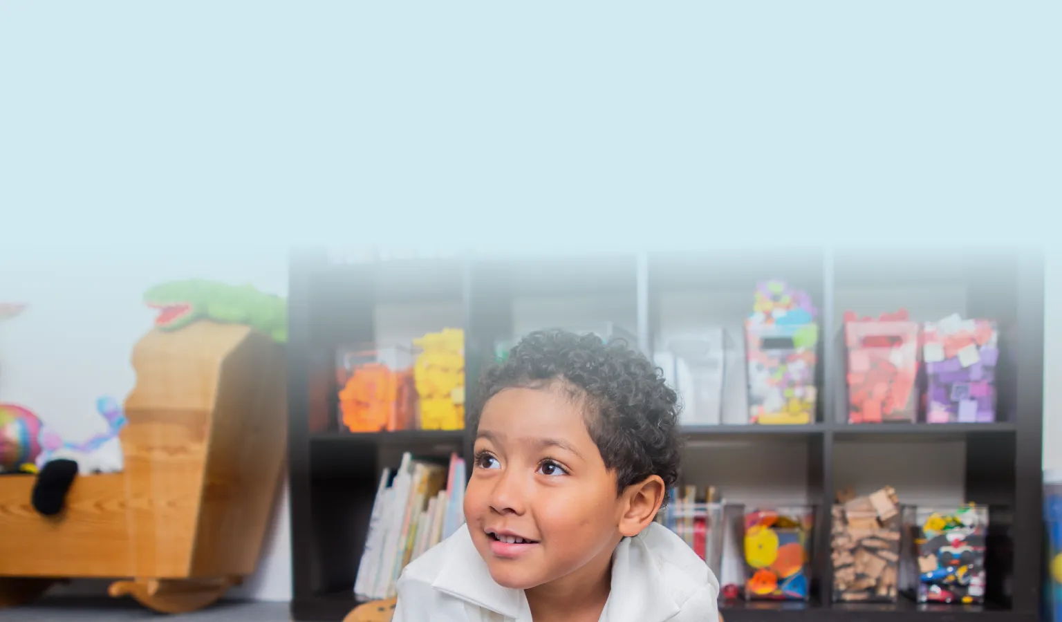A child with curly hair is playing on a playmat in a nursery. He looks up and to the left slightly, with an expression of hope on his face.