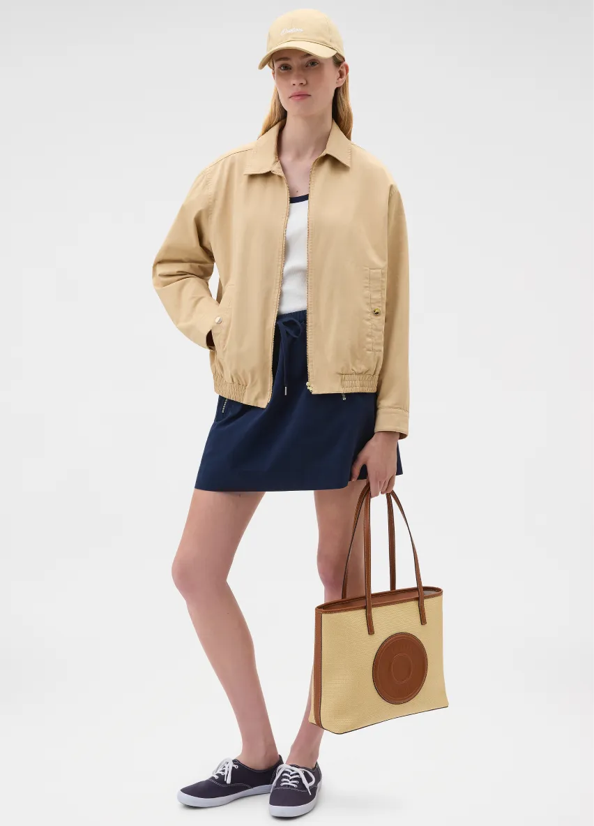 A woman in a beige jacket, navy skirt, and cap holding a tote bag.