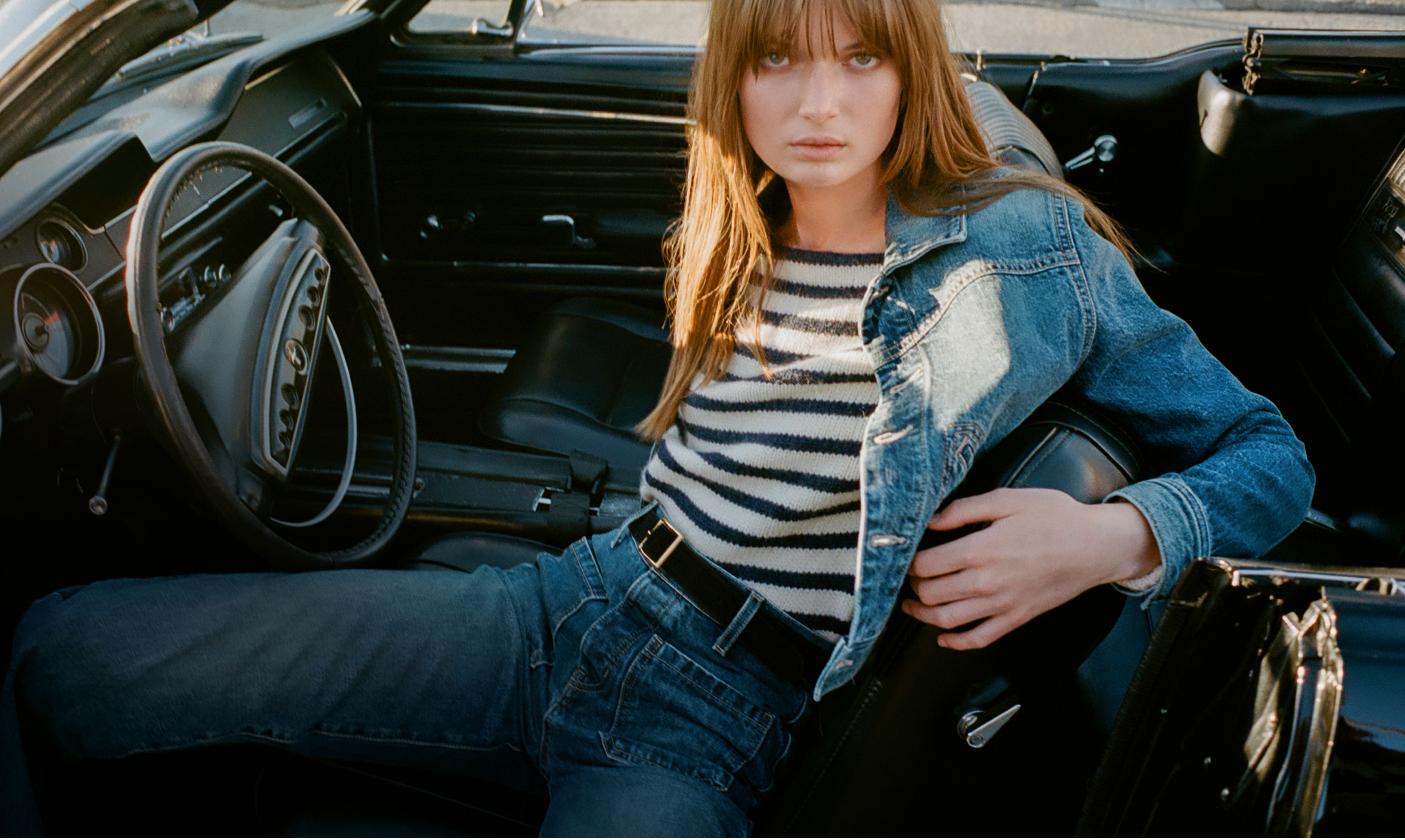 A person in a denim jacket and striped shirt sitting in a car.
