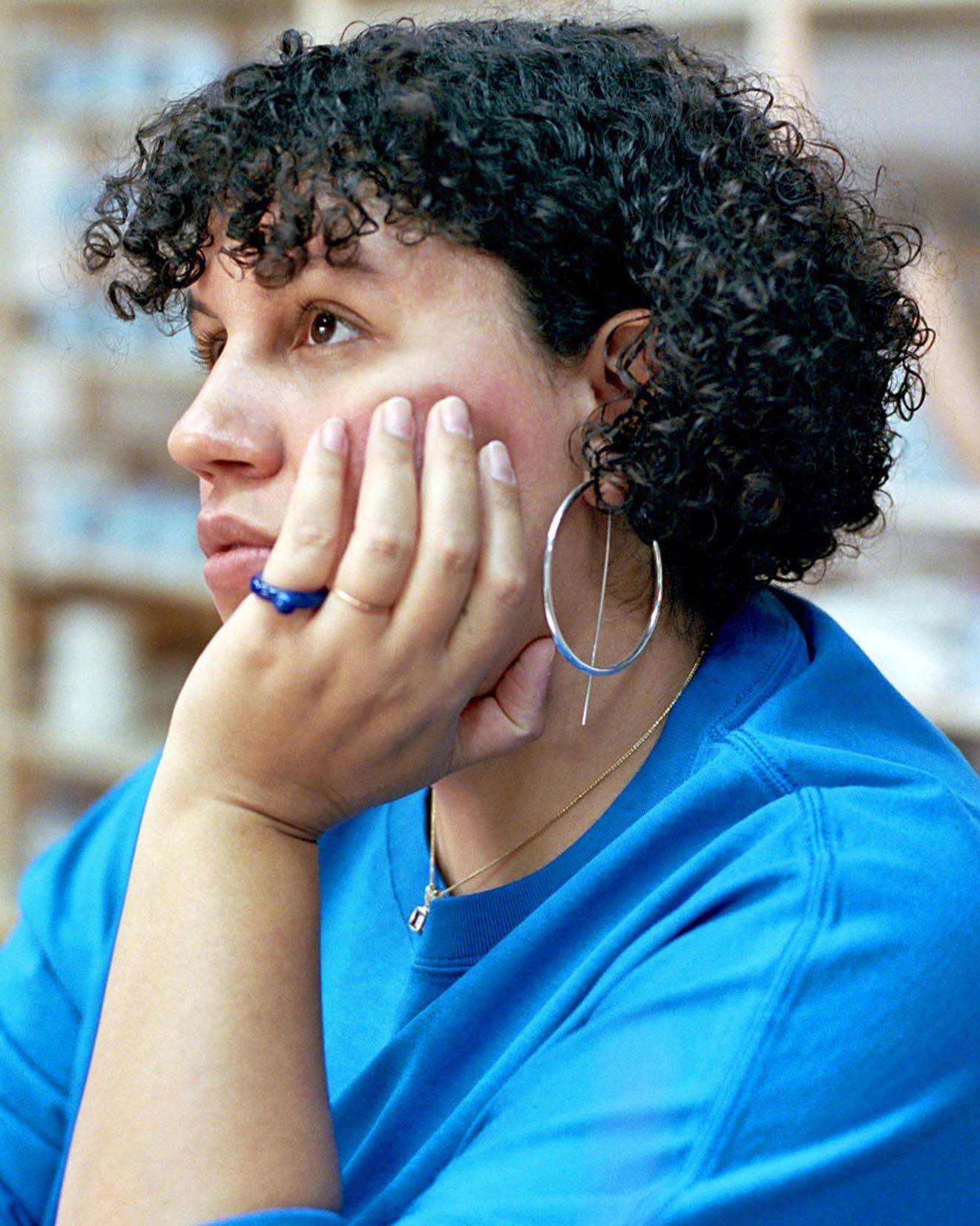 Close up image of Sara with her chin in her hand, wearing a blue glass ring, blue shirt, and oversized silver hoop earring. 
