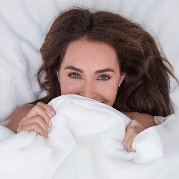 A person holding a beige Luxe™ brand throw over their shoulder, standing against a dark background.