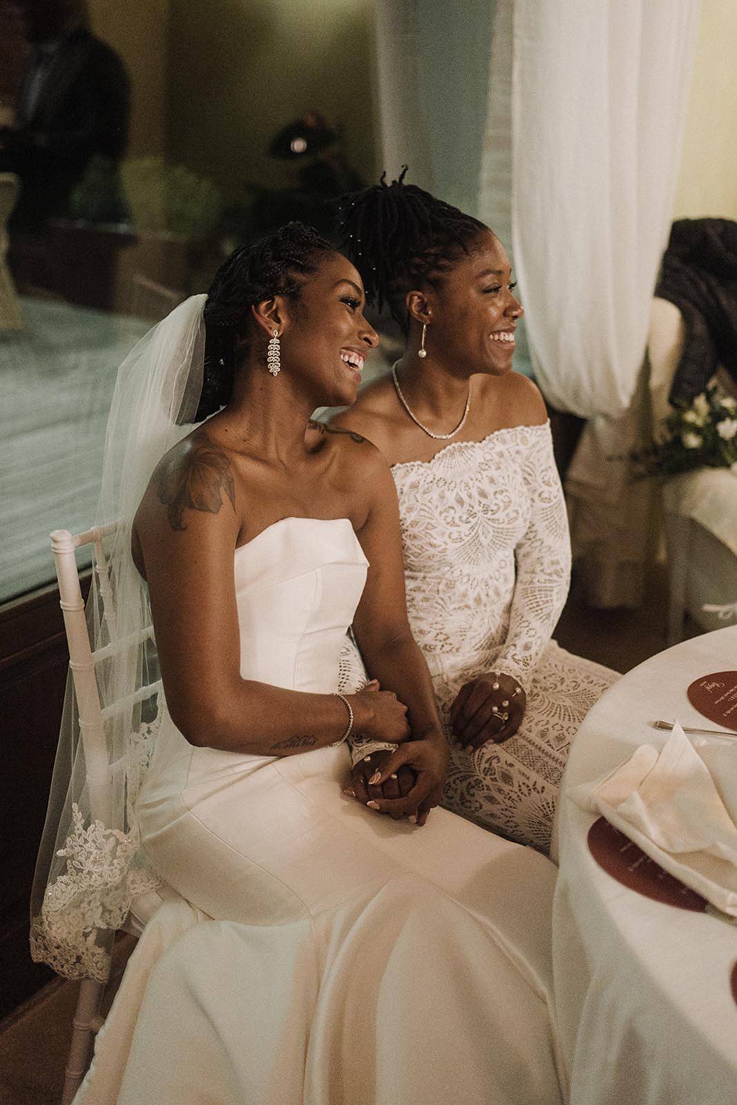 Two brides, beautifully adorned in white wedding gowns, sharing an intimate moment as they sit side by side