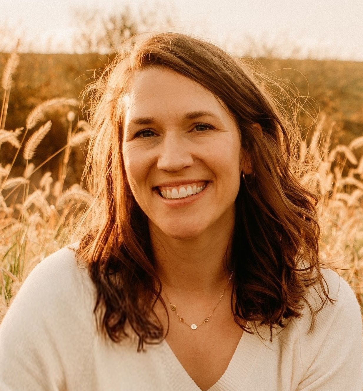 Smiling woman with wavy brown hair in a field during golden hour.
