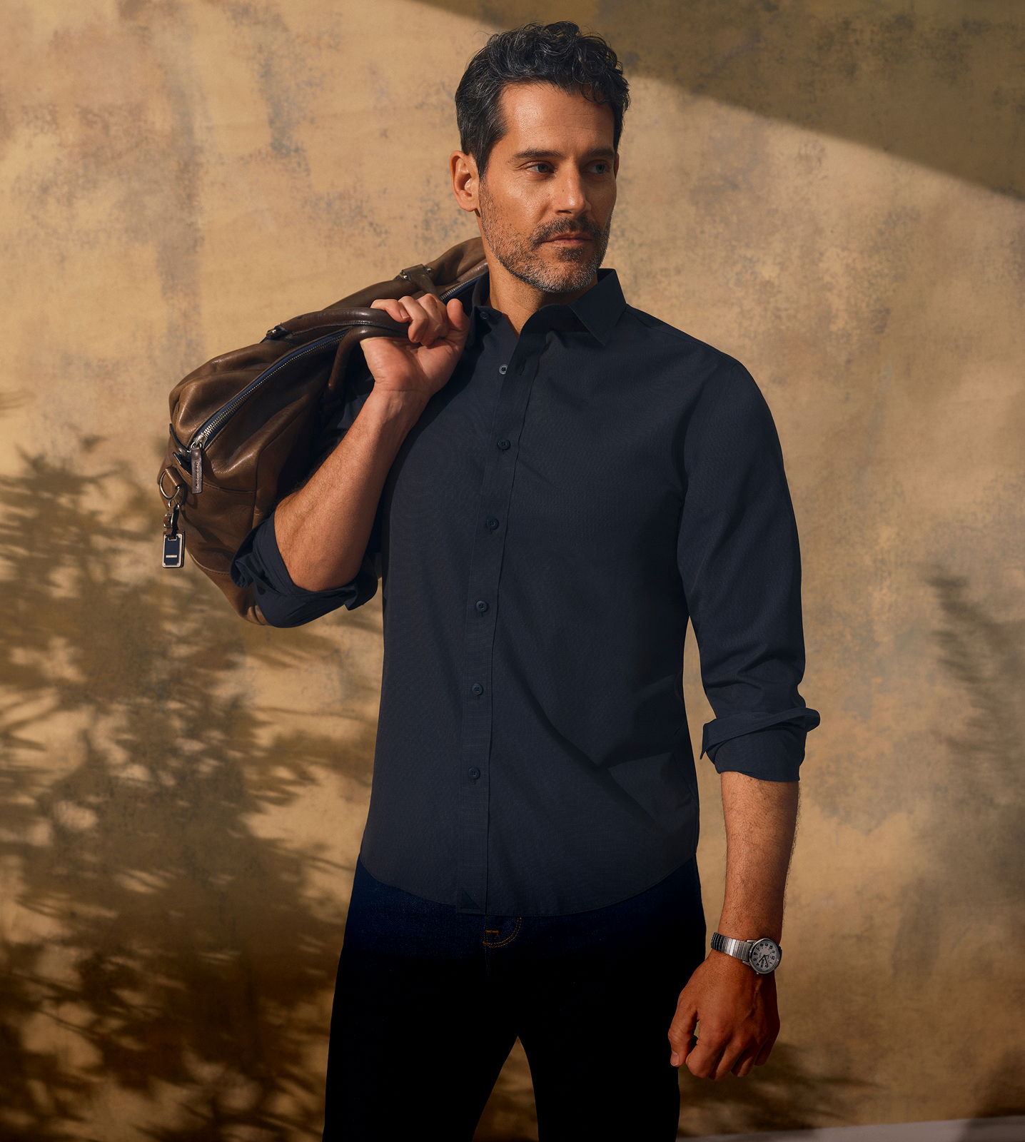 Man in a black shirt holding a jacket, standing against a textured background.