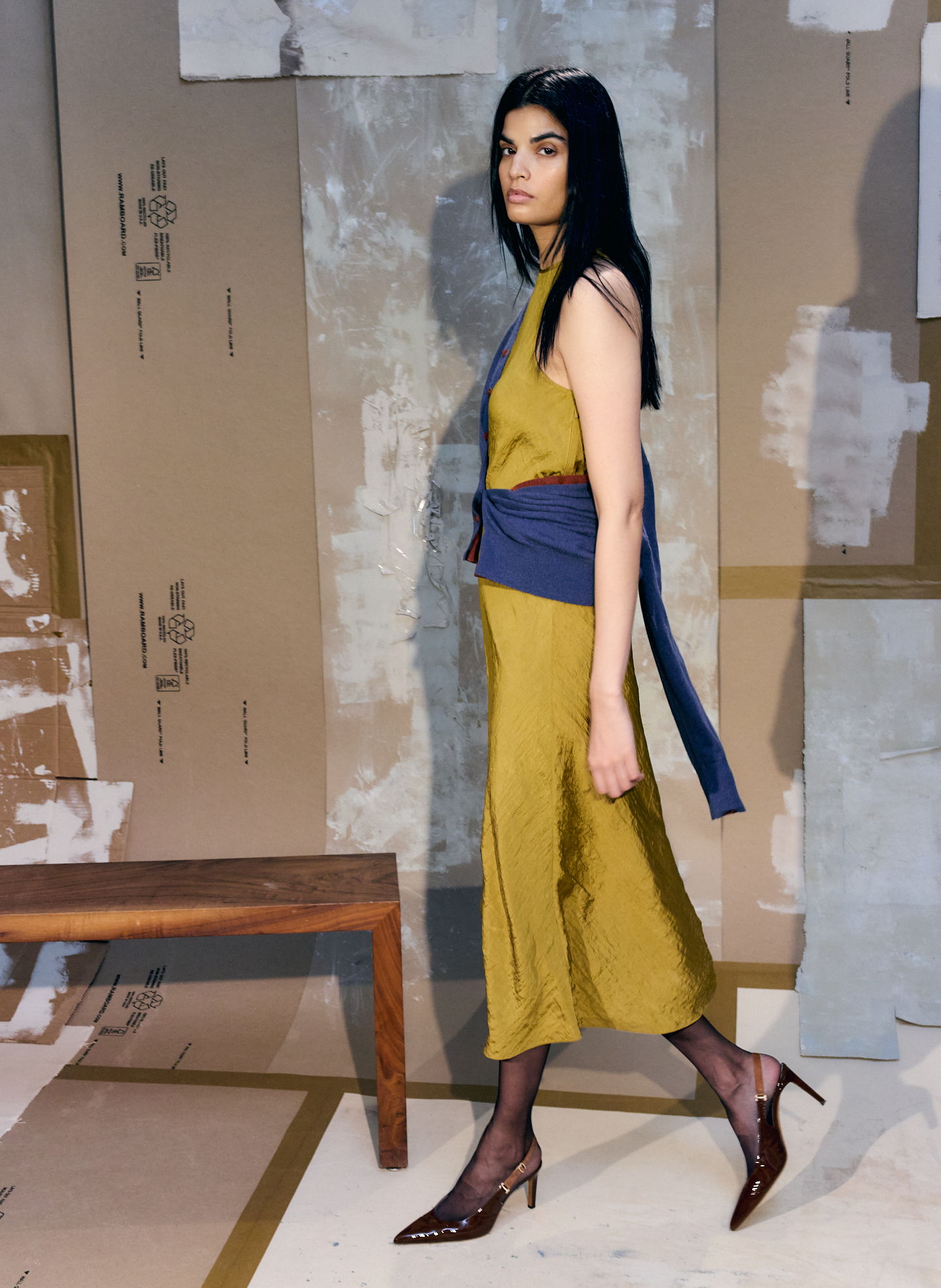 Model in a stylish outfit walking past a wooden bench and textured background.