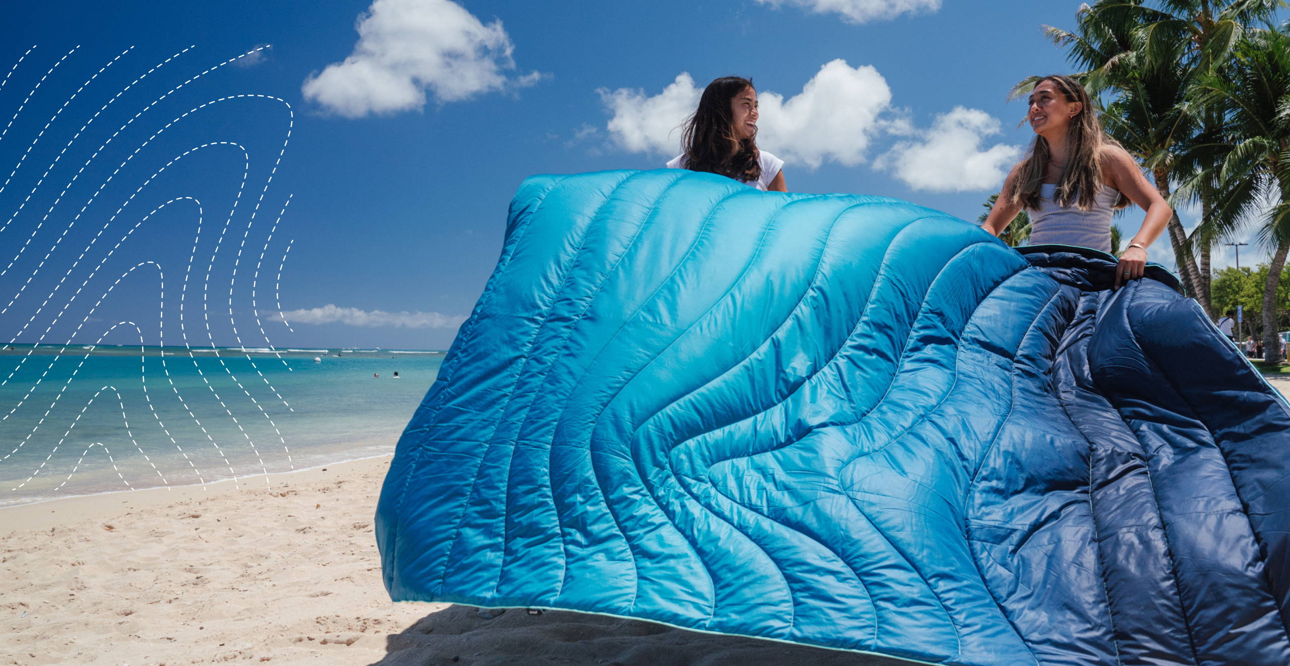 Two girls dust off a 2-person Ocean Fade beach blanket before laying it down on the sand.