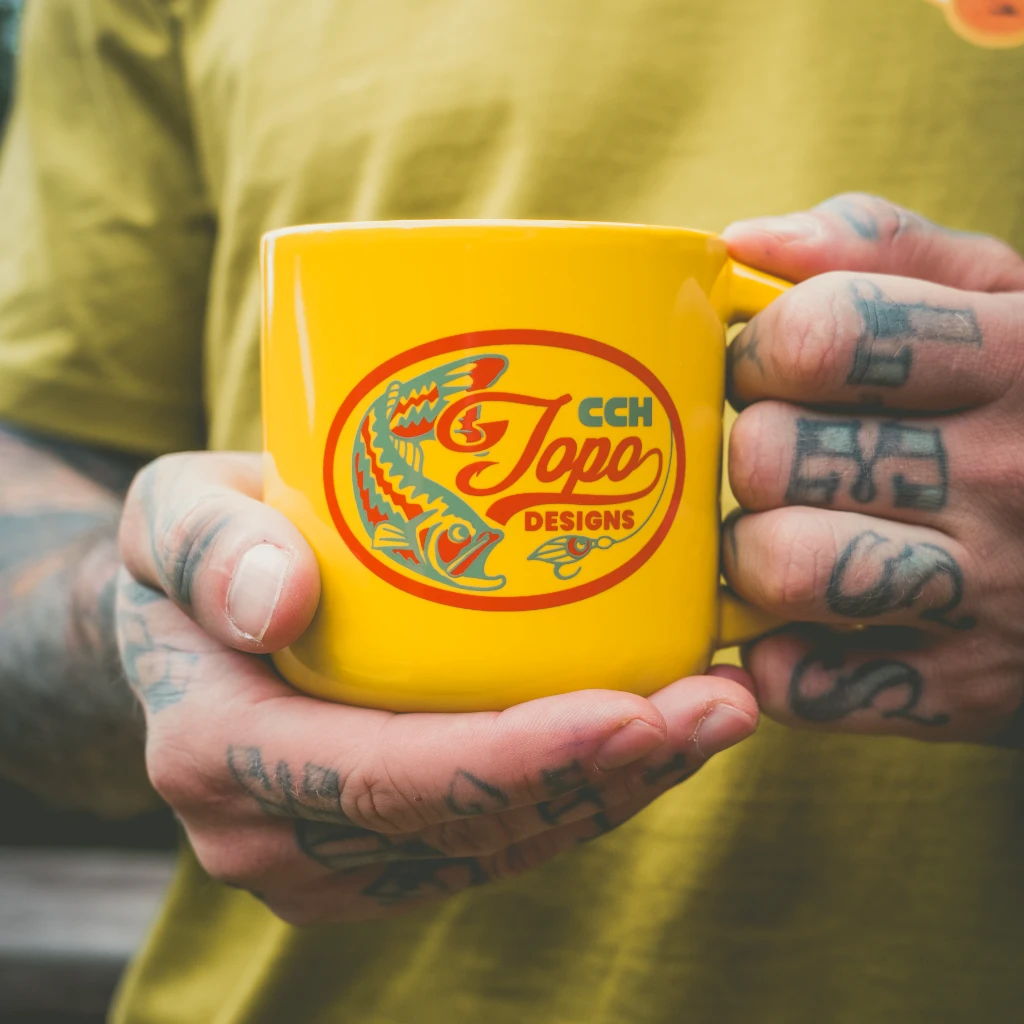 A person holding a yellow mug with a logo and tattoos on their hands.