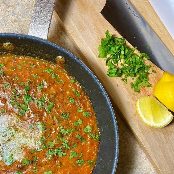 A cut-off bird’s eye view of a Misen Nonstick pan filled with an herby stew. Chopped herbs, lemon, and a Misen Chef’s Knife are visible to the side.