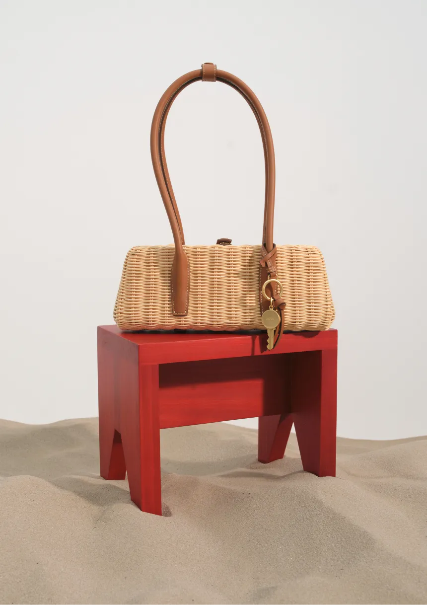 A woven handbag displayed on a red stool in sandy surroundings.