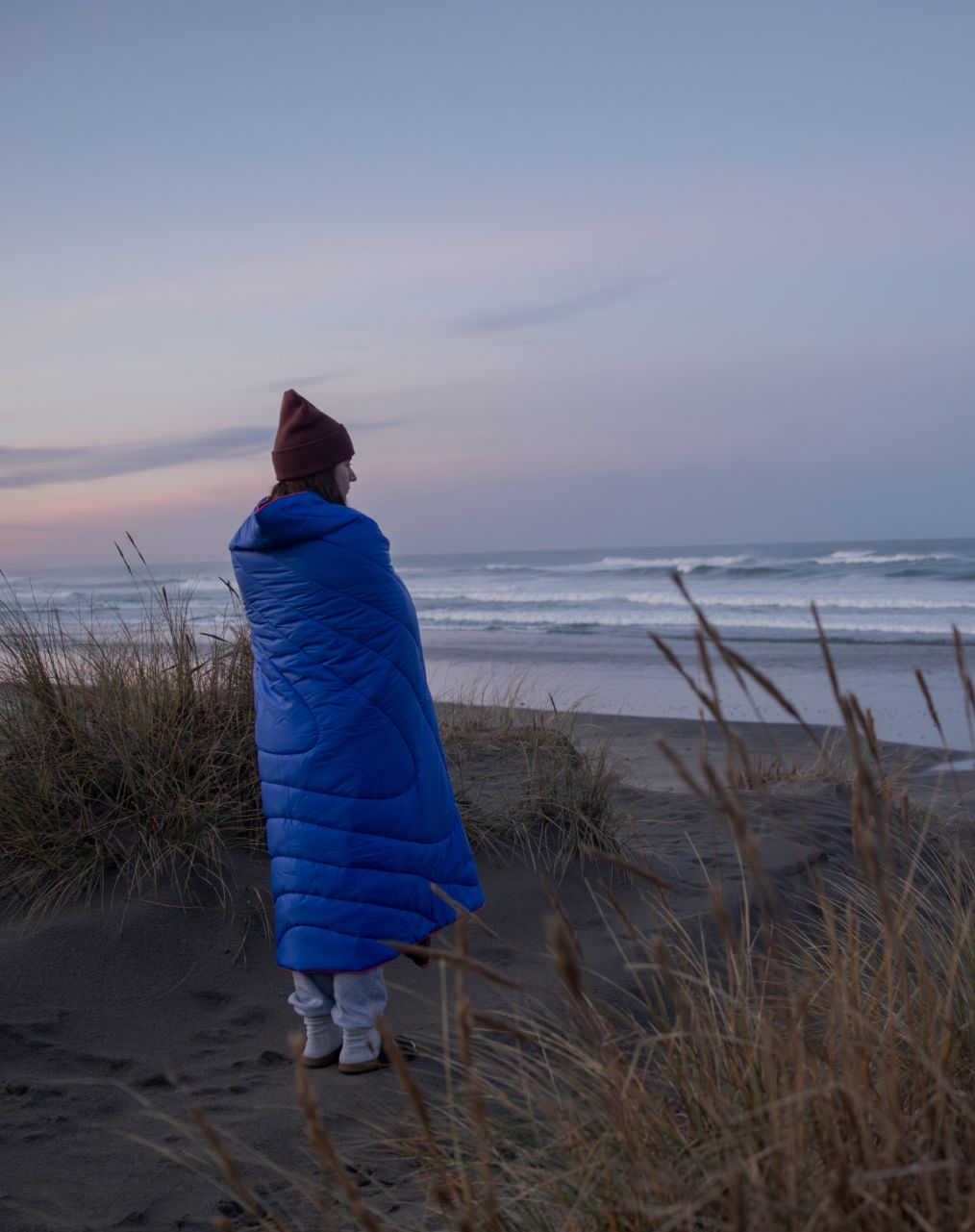 Person in a blue blanket standing on a beach at sunset.