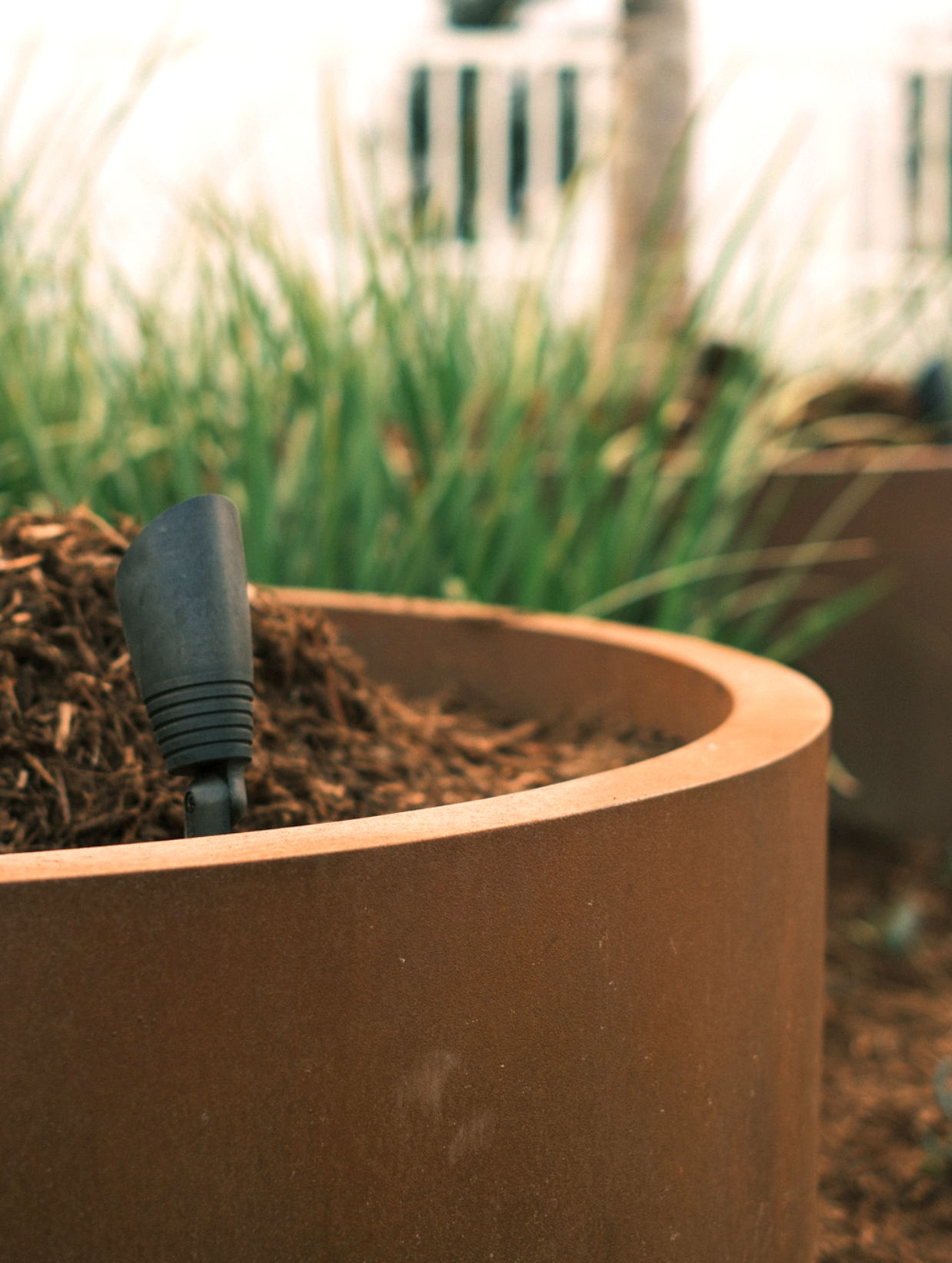 Planter with soil and a black gardening tool among green grass.