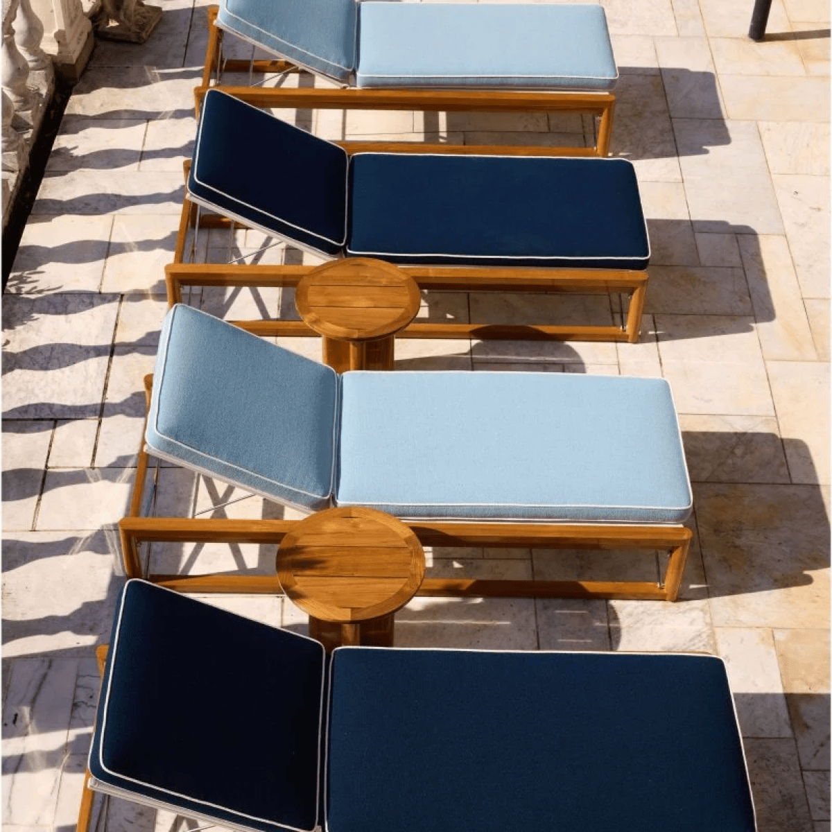 Teak poolside chaise lounges with blue cushions and small round side tables arranged for luxury pool patio design.