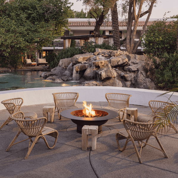 Outdoor fire pit seating area with woven lounge chairs, durable stone stools, and tropical landscaping beside a waterfall