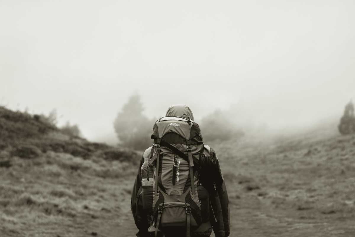 A person walking away from the camera with a backpack on a misty day , going up a hill with scrub on either side