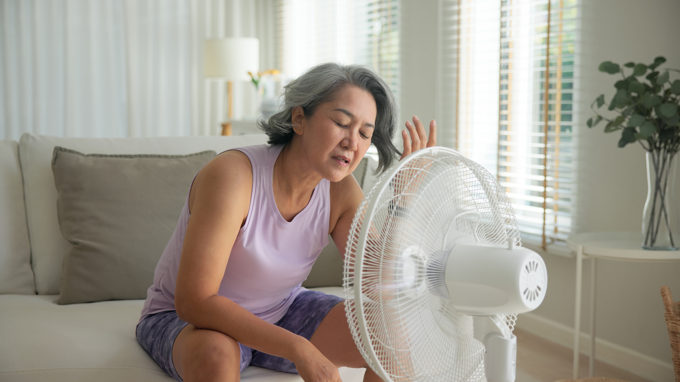 Woman sitting next to a fan, looking distressed in a bright room.