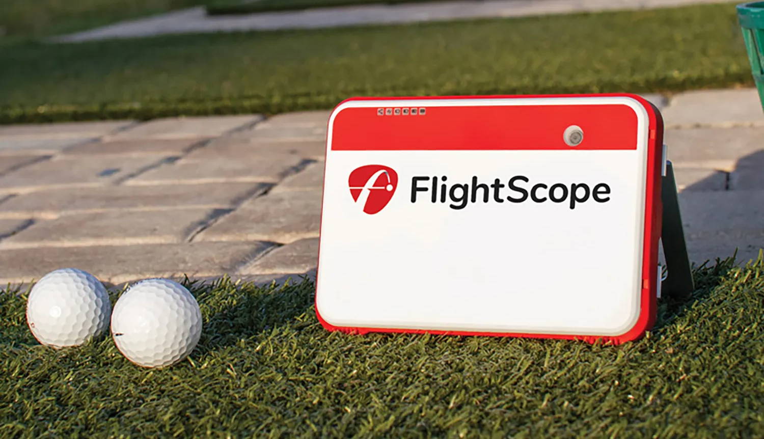 The FlightScope Mevo+ golf launch monitor sitting on the grass next to 2 golf balls and a stone patio