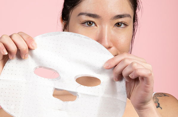 Woman holding a facial mask in front of her face, pink background.