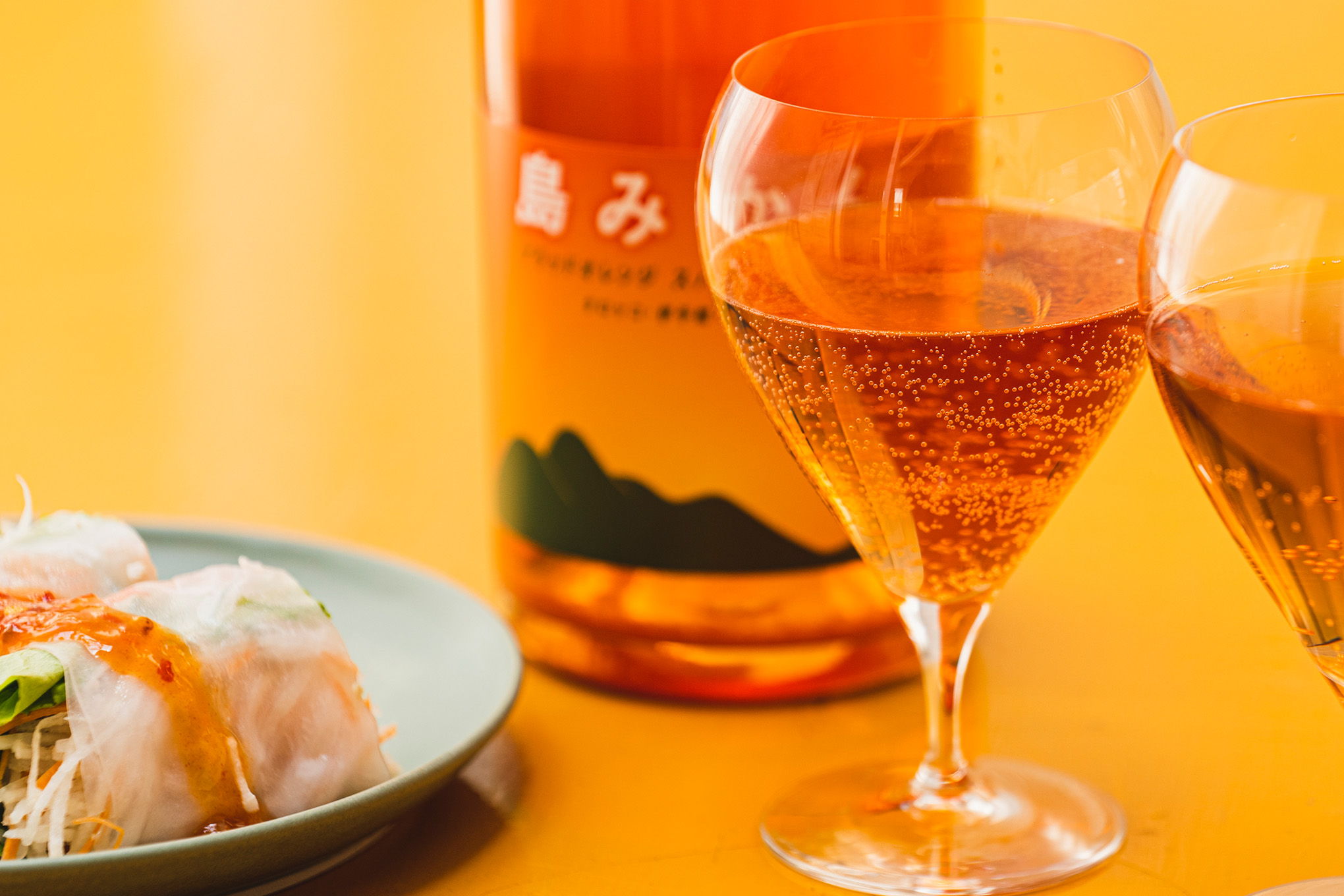 Two glasses of sparkling drink with sushi and a bottle on a yellow background.