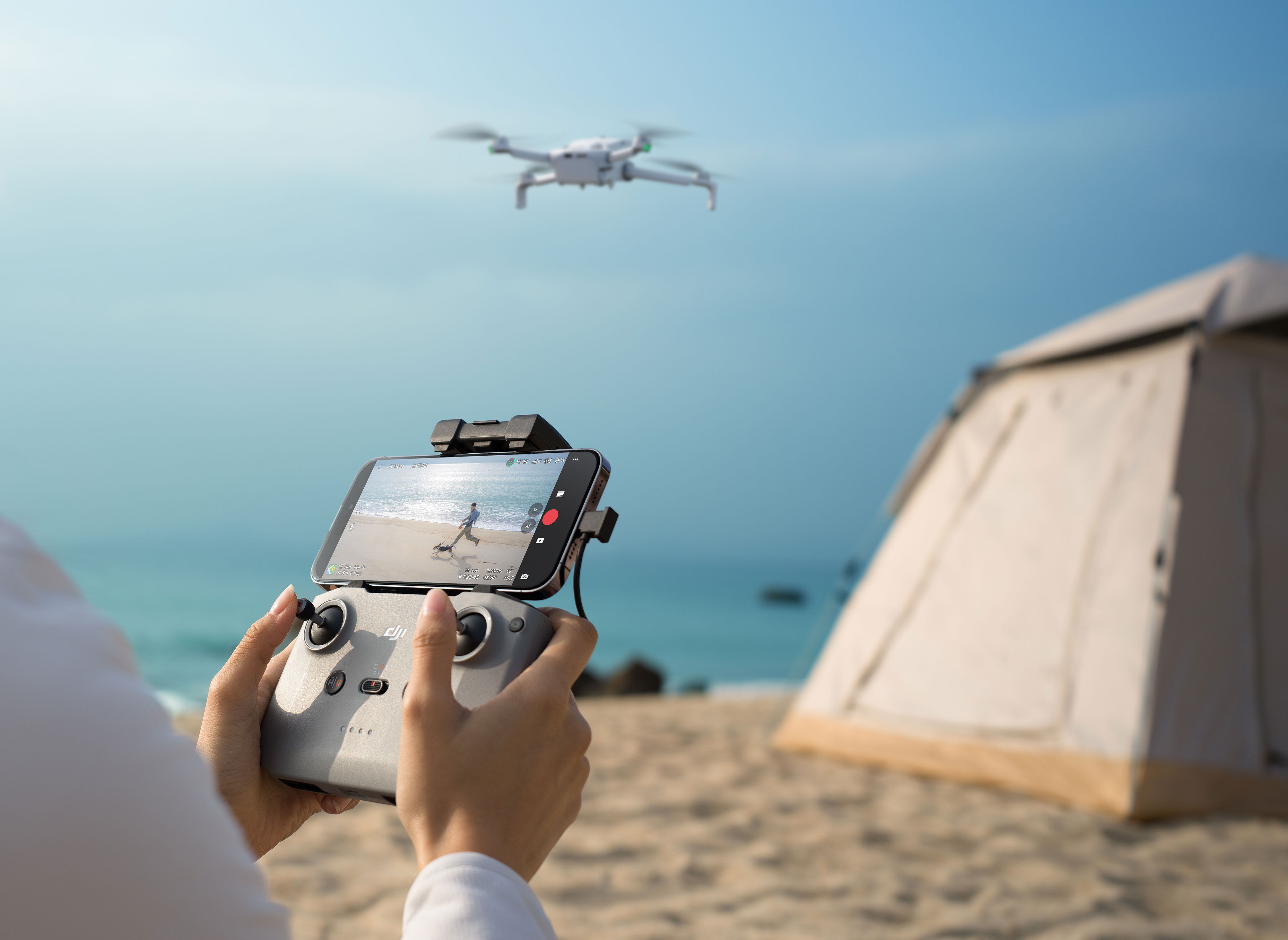 A person uses the DJI RC-N3 to fly the DJI Lito 1 near a tent on the beach.