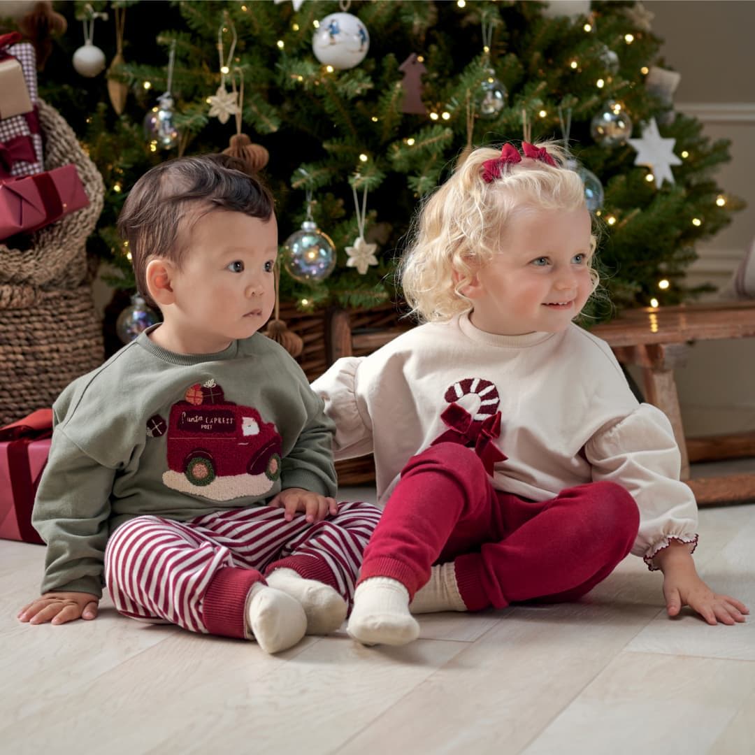 Two children sitting in front of a Christmas tree, wearing festive outfits.
