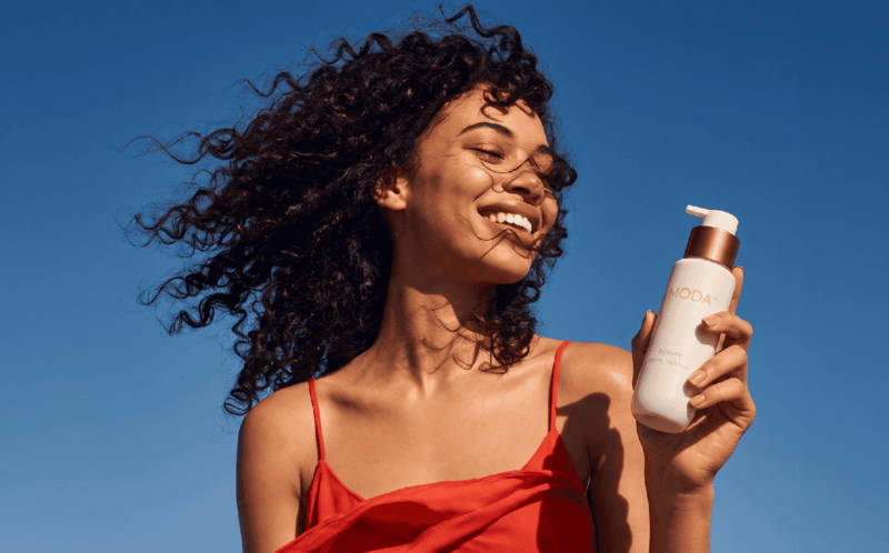 Woman with curly hair holding a MODA hair product bottle, blue sky background