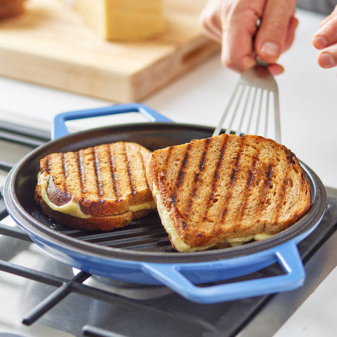 A hand uses a fish spatula to flip a grilled cheese sandwich with clear grill marks in a Blue Grill Lid on a stovetop.