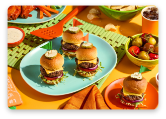 Three mini burgers on a colorful table with sides and condiments.