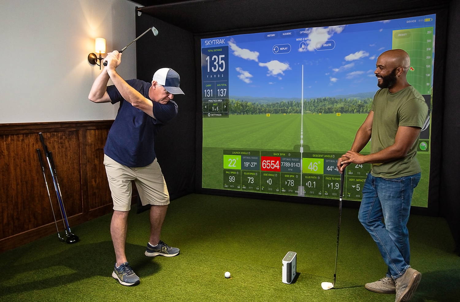 Two golfers, one swinging, one watching in an indoor golf simulator with a SkyTrak+ golf launch monitor in front of a large impact screen