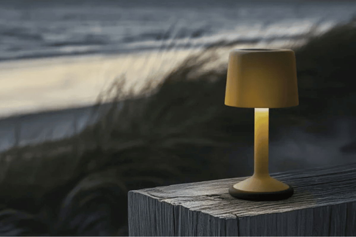 Small cordless outdoor table lamp in mustard yellow on a weathered wood post with dunes and ocean blurred in the background.