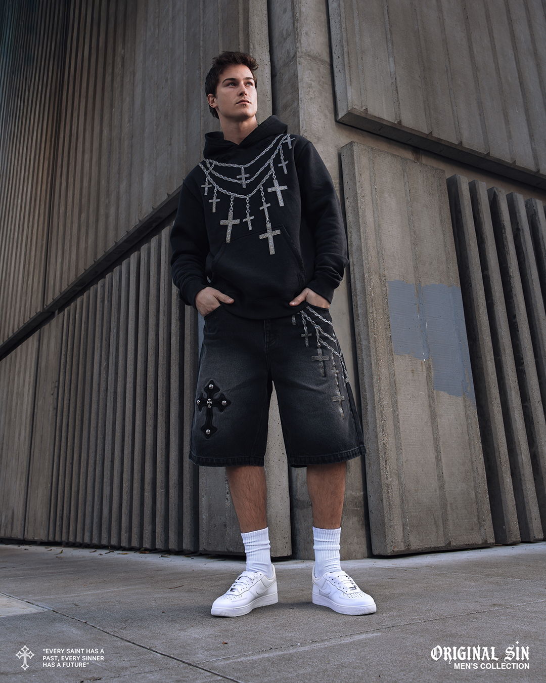 Young man in a black hoodie and shorts standing against a concrete wall.