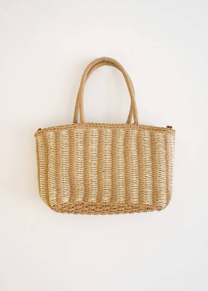 A wicker basket with a white and natural wicker striped pattern, handles and faux leather shoulder strap