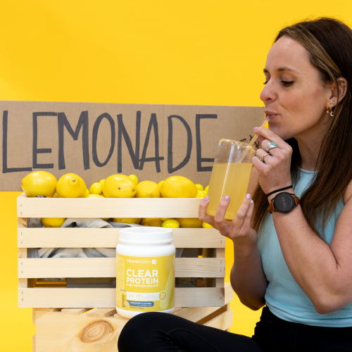 Woman sipping lemonade with lemons and a sign in the background.