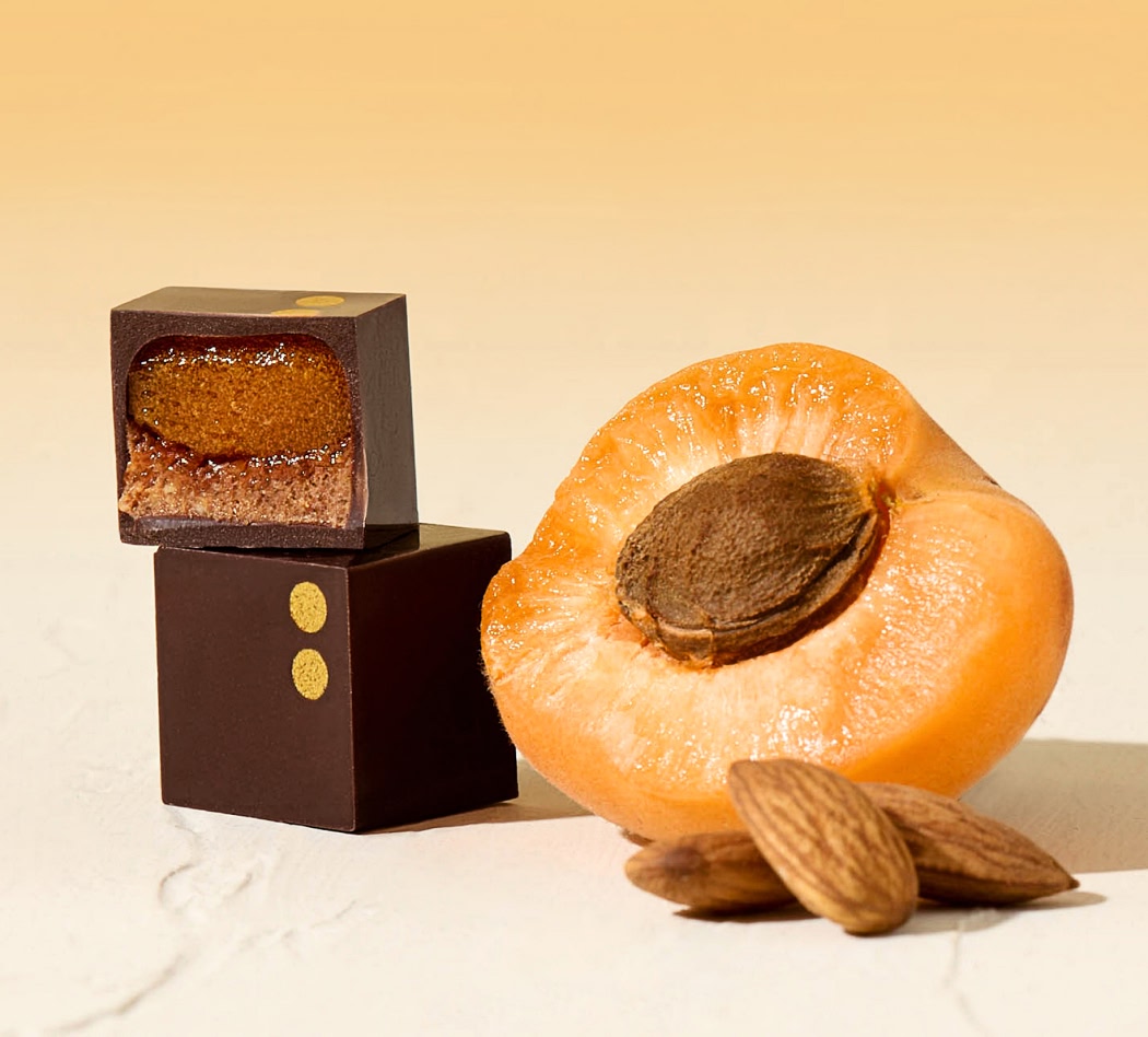 Chocolate cube beside a halved apricot and scattered almonds on a light background.