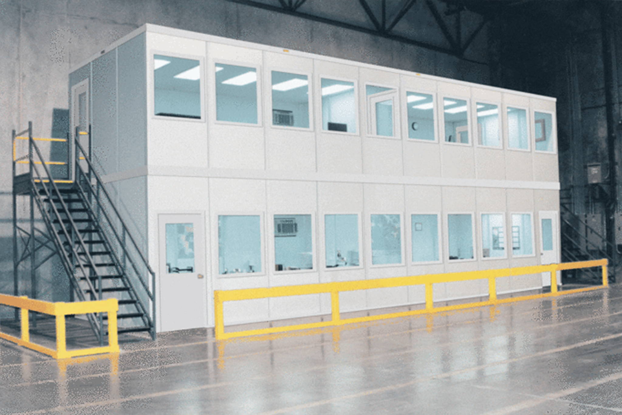 Two-story modular office installed in warehouse with mezzanine landing and stairs to access second floor office. Surrounded by yellow steel guard rails.
