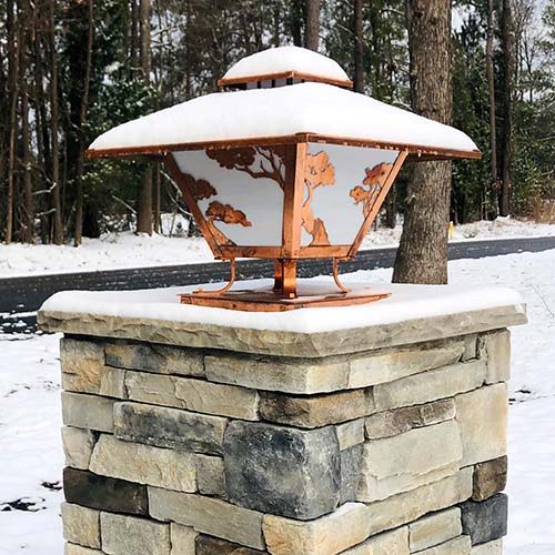 A two-story Arts & Crafts column light with Torrey pine artwork and a bright, raw copper finish. It's covered with a layer of snow.