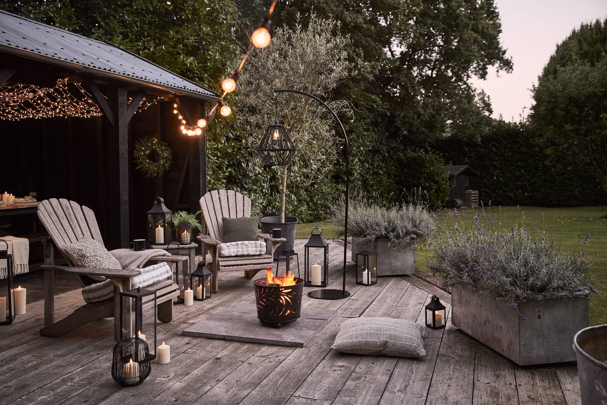 Un coin salon d'été extérieur avec des bougies et des lanternes LED, un coin brasero et des guirlandes guinguettes blanc chaud suspendues en hauteur.